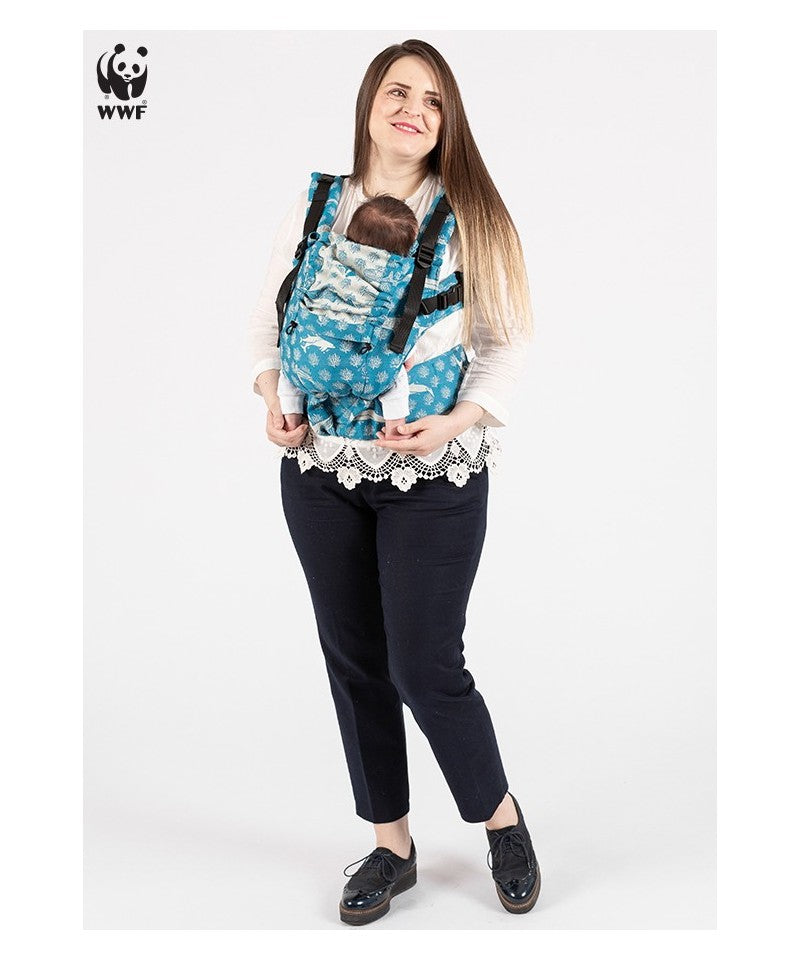 Mulher a usar a mochila porta-bebés ISARA The ONE, ajustada para um recém-nascido. A mochila é azul com um padrão de espécies marinhas. A mãe sorri e segura o bebé junto ao peito.