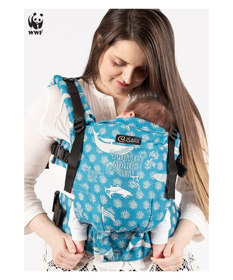 Mulher a usar a mochila porta-bebés ISARA The ONE, ajustada para um recém-nascido. A mochila é azul com um padrão de espécies marinhas. A mãe sorri e segura o bebé junto ao peito.