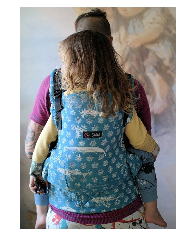 Vista traseira de uma mulher a usar a mochila porta-bebés ISARA The ONE, com uma criança nas costas. O design Marine Life é claramente visível e a mochila está ajustada para um ajuste seguro e confortável.