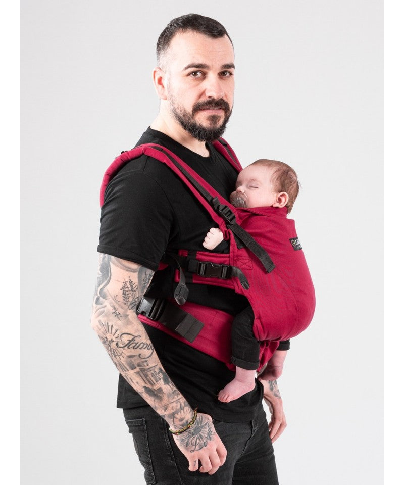 Pai com tatuagens de pé com um porta-bebés vermelho ISARA The ONE, visível de corpo inteiro.