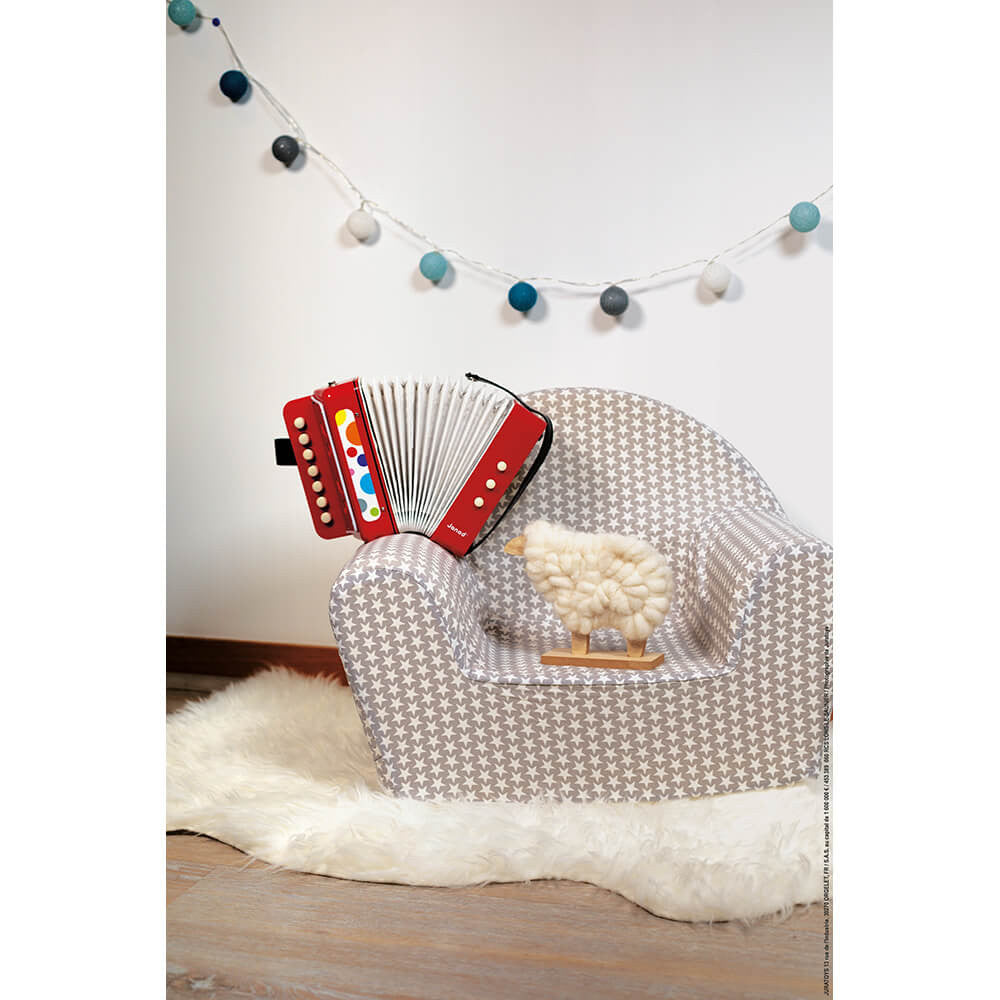 Acordeão Janod Confetti vermelho pousado numa cadeira infantil, com um brinquedo de ovelha.