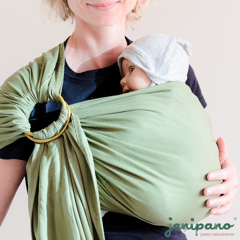 Uma mulher usando uma t-shirt preta segura um bebé com um chapéu cinza, transportado num sling de argolas verde da marca Jenipano. O sling está bem ajustado e o bebé parece confortável, próximo do peito da mulher.