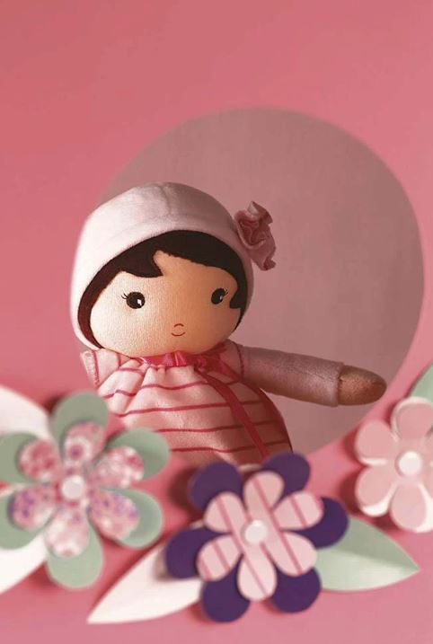 Close-up da boneca Kaloo A minha primeira boneca Rosa, com fundo rosa artístico e flores de papel, focando no rosto e gorro.