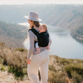 Vista traseira de uma mulher a carregar um bebé na Mochila Neobulle MyNeo, com uma paisagem natural ao fundo.