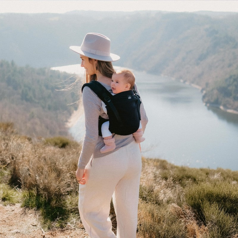 Vista traseira de uma mulher a carregar um bebé na Mochila Neobulle MyNeo, com uma paisagem natural ao fundo.