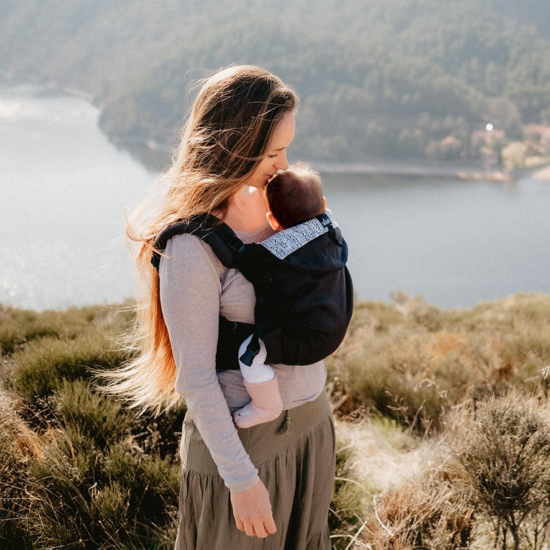 Mulher usando a Mochila Neobulle Ajustável ToddlerSize Ébène ao ar livre, com o bebé carregado à frente, em cenário natural.