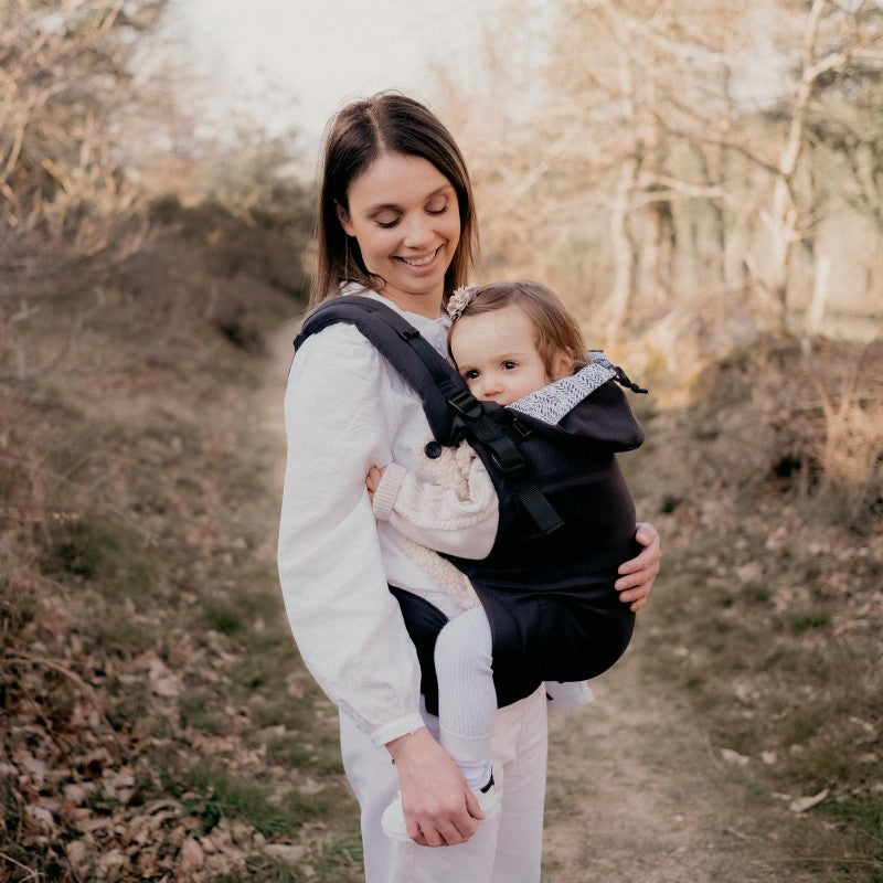 Mulher caminhando ao ar livre usando a Mochila Neobulle Ajustável ToddlerSize Ébène, com o bebé carregado à frente, ambos sorridentes.