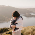 Imagem de uma mãe a usar a Mochila Neobulle com o bebé na posição frontal, destacando a versatilidade do porta-bebés em ambiente ao ar livre.