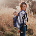 Mãe a usar a Mochila Neobulle Néo + azul, carregando um bebé nas costas enquanto caminha num campo.