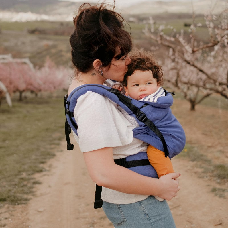 Mãe a usar a Mochila Neobulle Néo + azul, carregando um bebé pequeno na posição frontal durante um passeio.