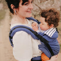 Mãe sorridente a usar a Mochila Neobulle Néo + azul, carregando o seu bebé na posição frontal, com natureza ao fundo.