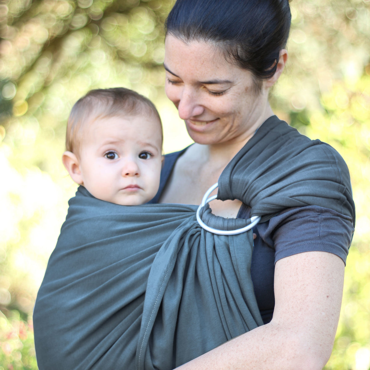Uma mãe sorridente carrega o seu bebé num sling de argolas cinzento, feito de jersey macio. O bebé está confortavelmente apoiado e olha para a frente, enquanto a mãe segura o sling de forma segura e ajustada.