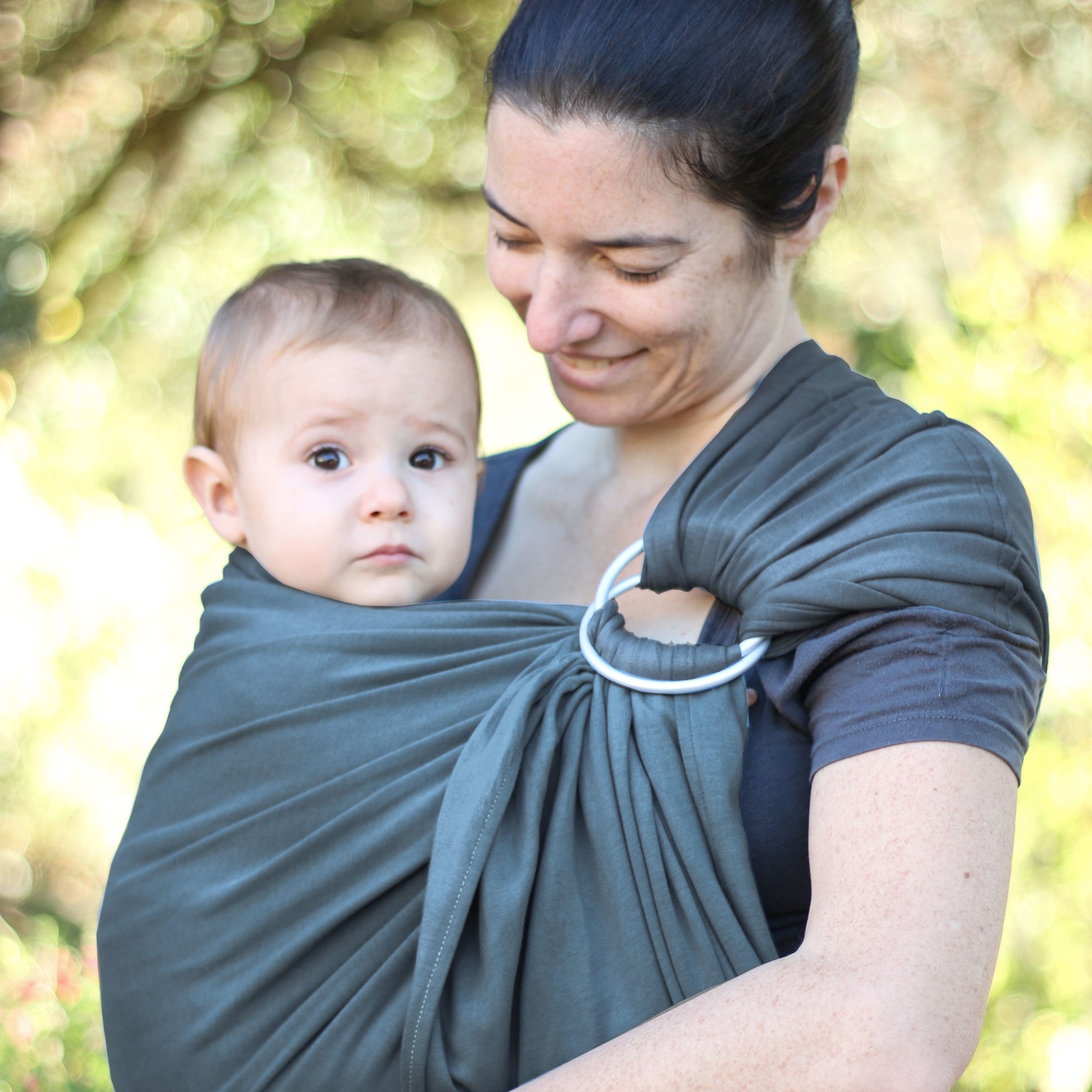 Uma mãe sorridente carrega o seu bebé num sling de argolas cinzento, feito de jersey macio. O bebé está confortavelmente apoiado e olha para a frente, enquanto a mãe segura o sling de forma segura e ajustada.