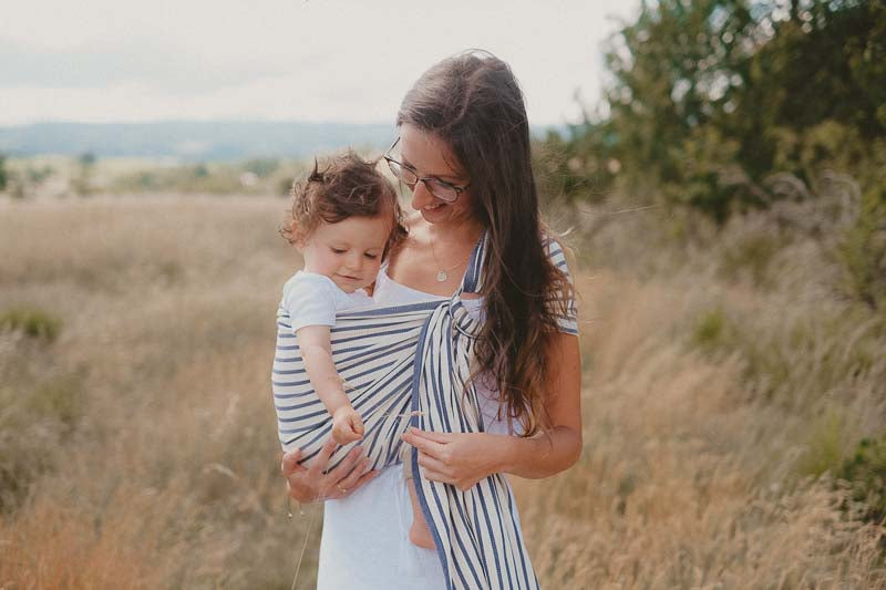Uma mãe segura o seu bebé num campo aberto, utilizando um Sling de Argolas Neobulle Malo. O pano é listrado e ajusta-se perfeitamente ao corpo da mãe e do bebé, oferecendo suporte e conforto.