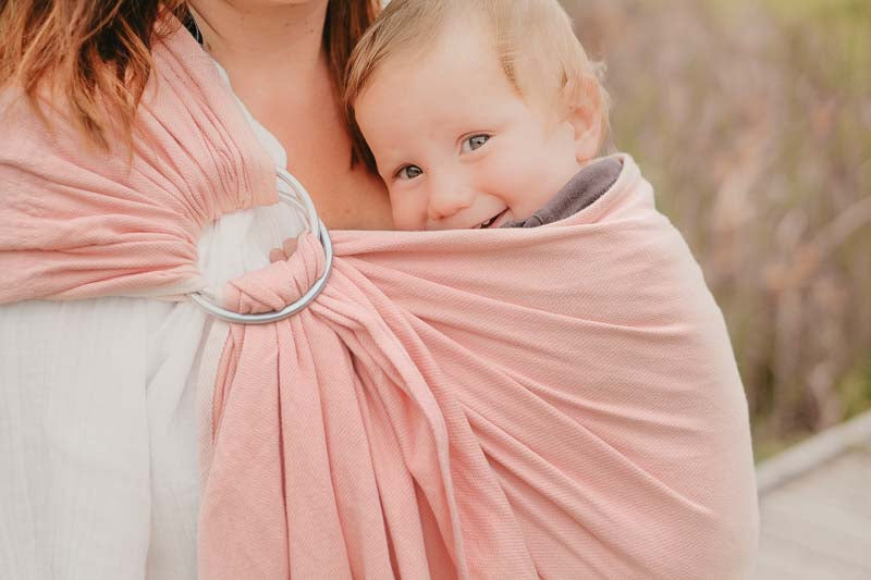 Mãe carregando bebé num sling de argolas rosa, ambos sorrindo, com fundo natural desfocado.