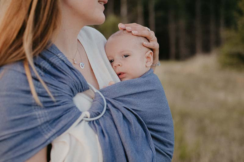 Mulher a usar um sling de argolas Neobulle Bleu Cobalt, com um bebé aninhado confortavelmente.