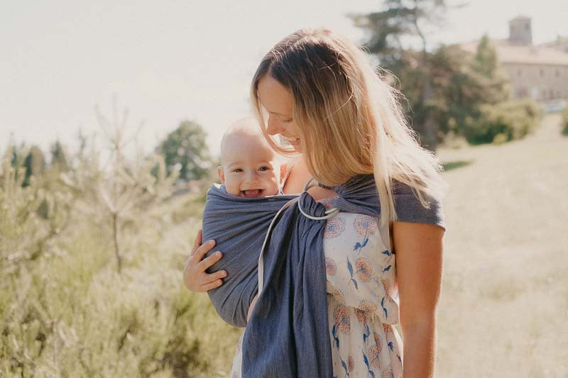 Mãe a caminhar ao ar livre com o seu bebé no sling de argolas Neobulle Bleu Cobalt, evidenciando a praticidade do produto.