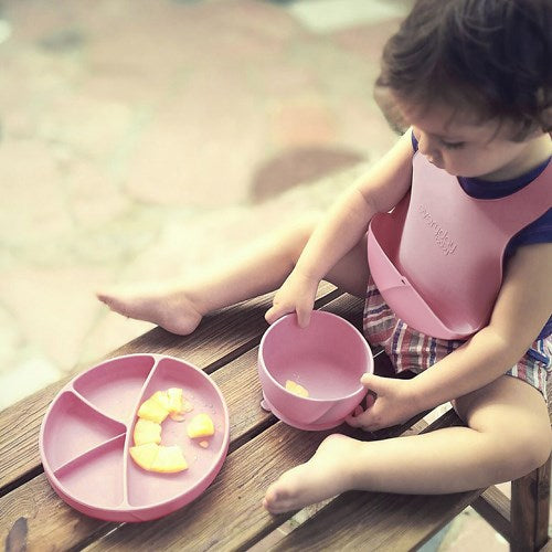 Criança pequena sentada num banco de madeira ao ar livre, a brincar com um prato de silicone rosa e uma taça separada com pedaços de fruta.