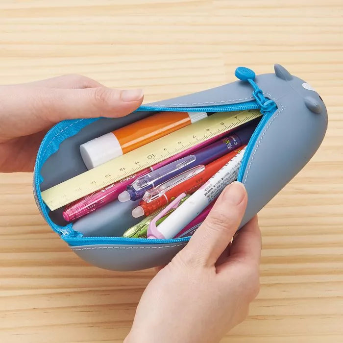 Mãos a segurar um estojo de silicone cinzento aberto, cheio de canetas, lápis e uma régua de 15 cm, mostrando o interior espaçoso.