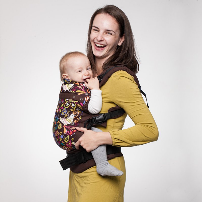 Uma mãe sorridente usando a Mochila Liliputi com um padrão vibrante de pássaros, carregando um bebé feliz. A mochila tem um painel fixo de mais de 60cm, proporcionando suporte ergonómico e confortável.