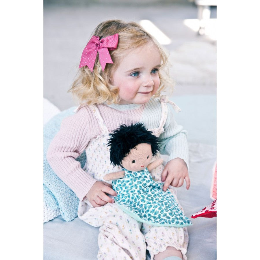 Criança pequena de cabelo loiro sentada a segurar a boneca Ari da Lilliputiens, que tem cabelo escuro e um macacão estampado.