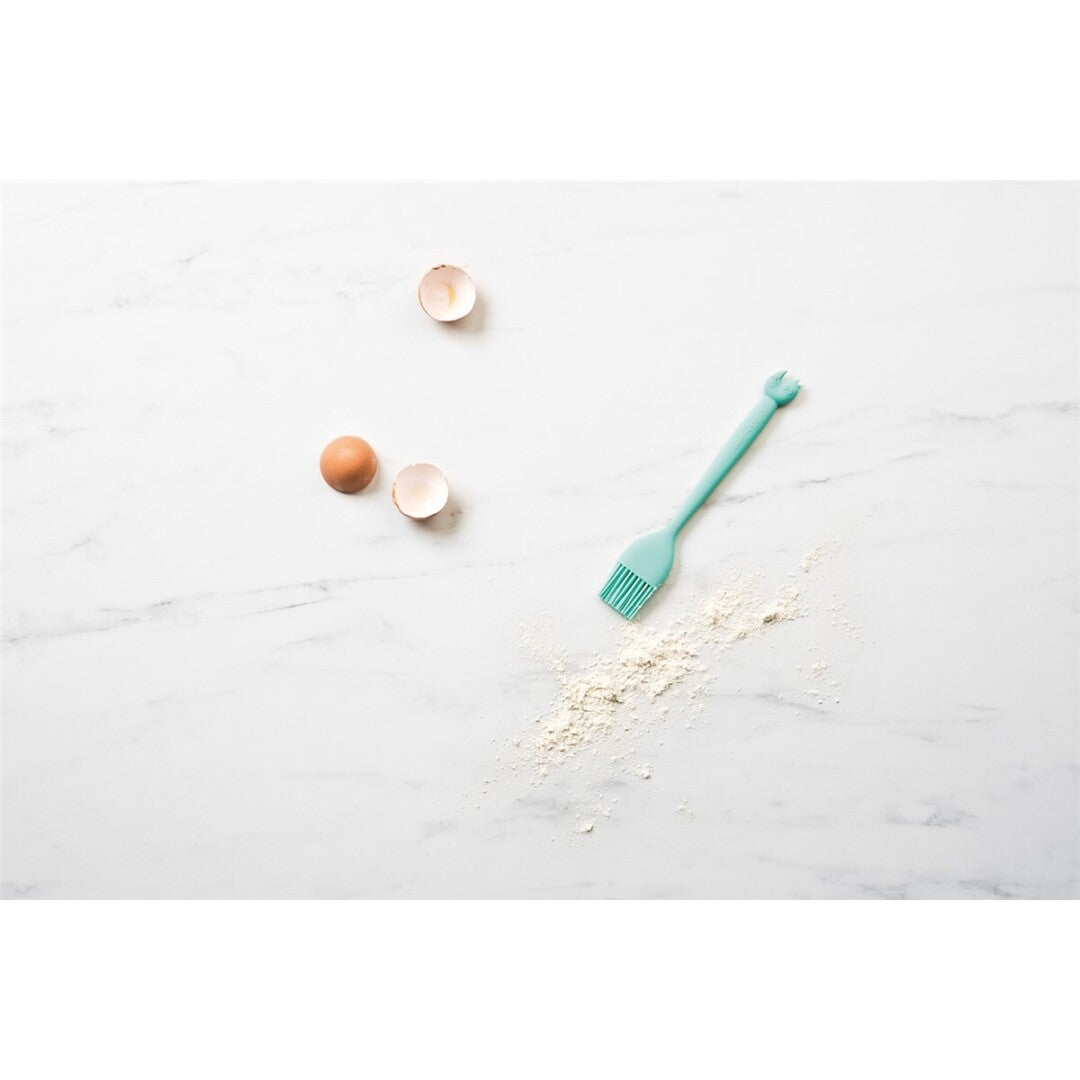 Pincel de cozinha Lilliputiens Little Chef em silicone azul-água, deitado sobre mármore com farinha e cascas de ovo, sugerindo uso culinário.