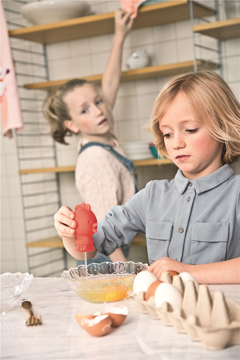 Criança loira a usar o separador de ovos Lilliputiens (forma de galinha) para separar a gema de ovo numa tigela de vidro, em atividade de culinária.