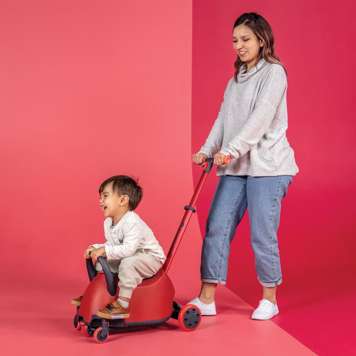 Mãe empurra o filho no Glider Luna, que está no modo passeio com pega de controlo, num cenário com fundo rosa e azul.