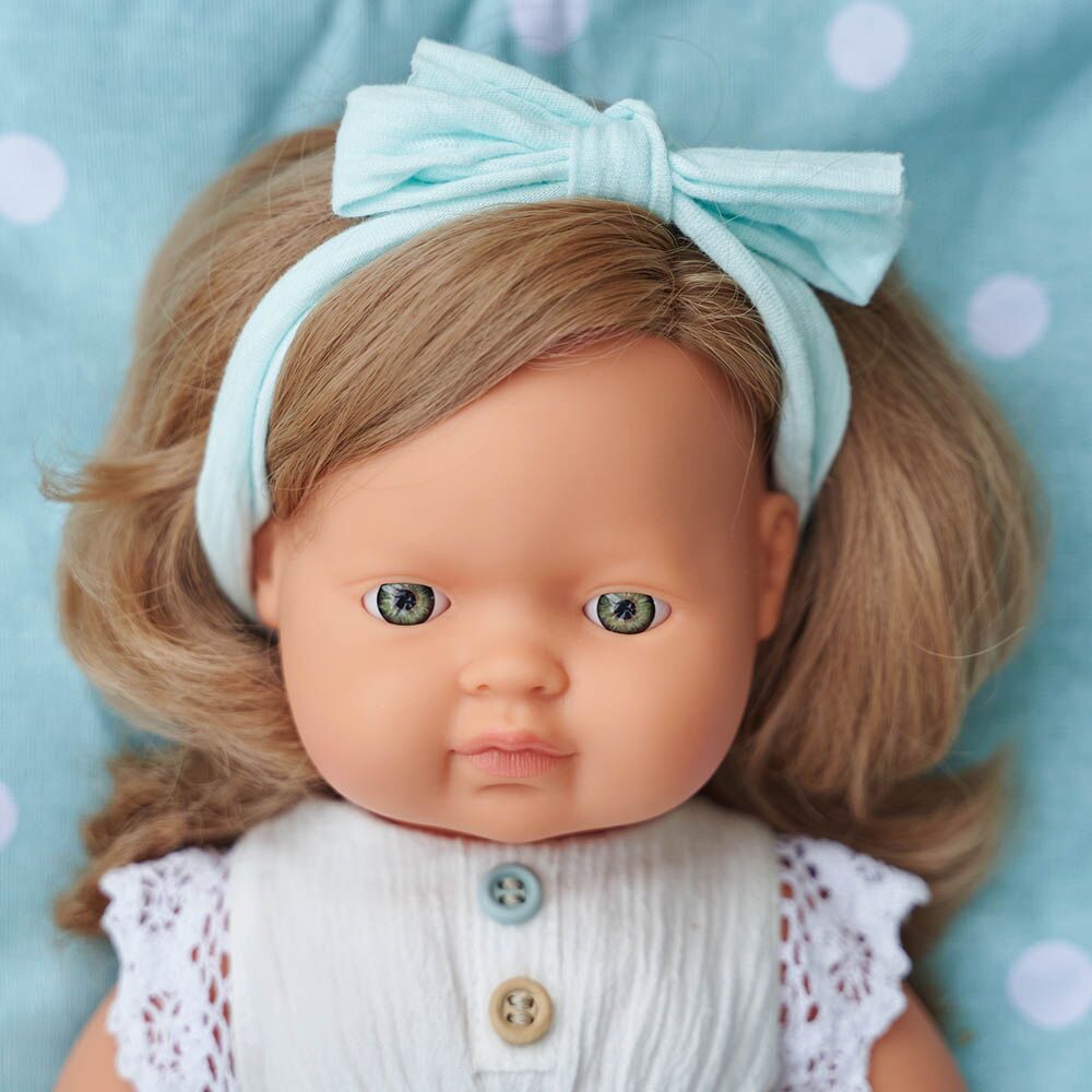 Close-up do rosto de uma boneca Miniland com cabelo louro, laço azul-claro e vestido branco com botões, sobre fundo de bolinhas.