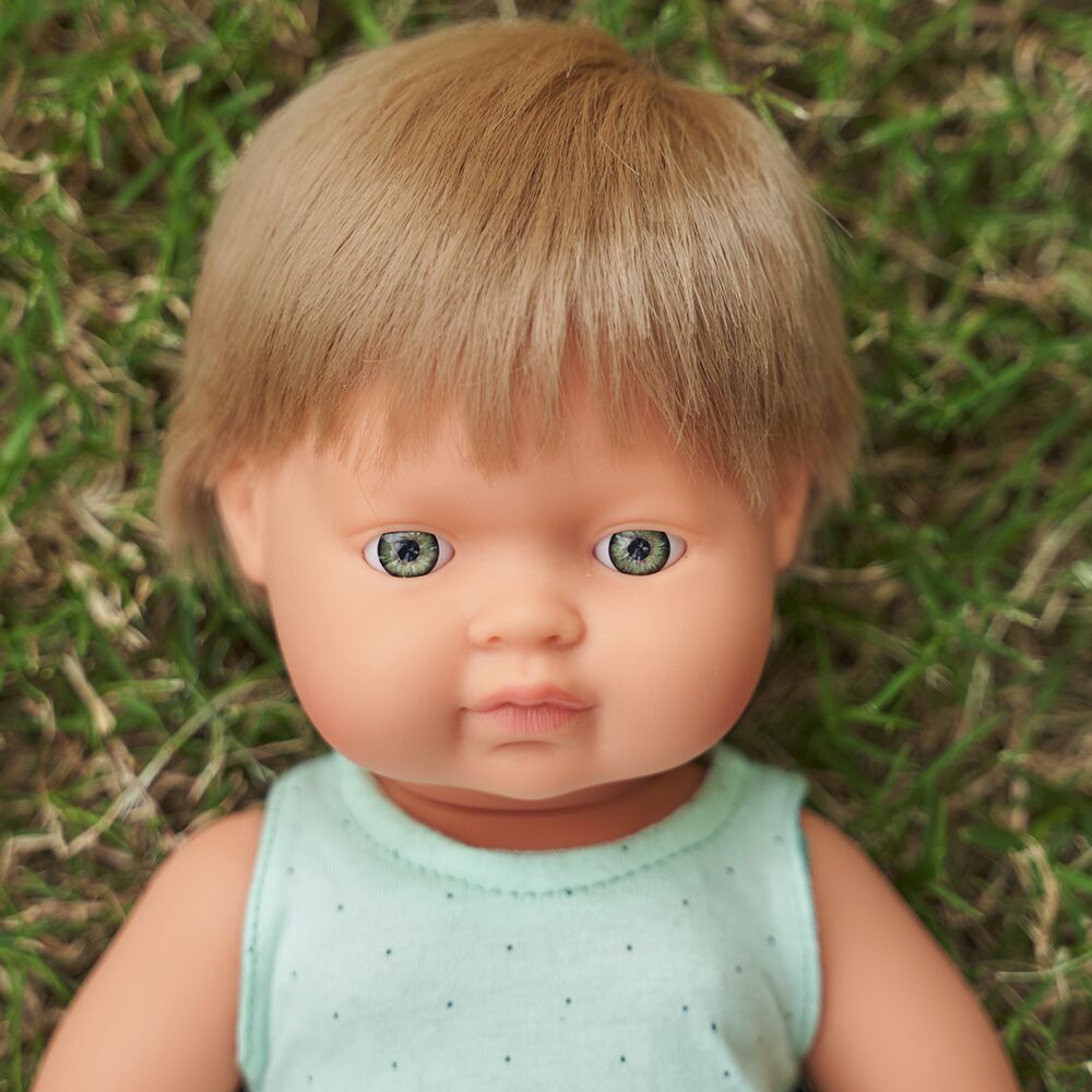 Close-up do rosto de um boneco bebé Miniland com cabelo louro e olhos verdes, deitado sobre relva verde.