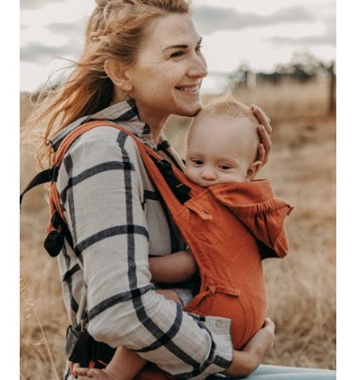 Uma mãe sorridente usando uma mochila Fidella Fusion Evolutiva Cubic Lines Rustred, com o seu bebé confortável e seguro na posição frontal, em um ambiente externo natural.