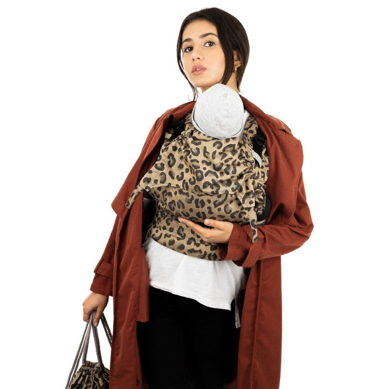 Mulher com cabelo castanho, vestida com um casaco castanho e camisa branca, a carregar um bebé numa mochila Fidella Fusion Evolutiva Leopard Gold.