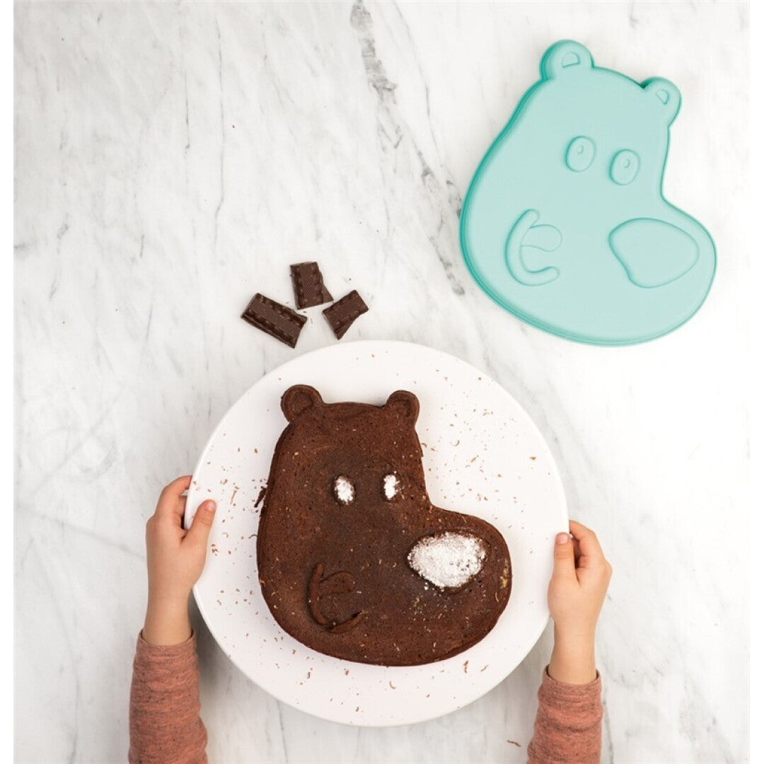 Bolo de chocolate em forma de cabeça de hipopótamo César finalizado, servido num prato branco, com o molde de silicone azul ao lado. Mãos de criança seguram o prato.