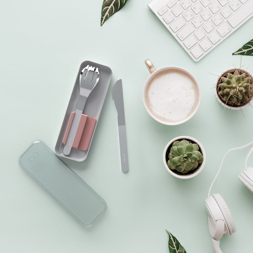 Talheres Monbento em estojo cinza-claro e estojo de transporte verde-água, dispostos numa secretária com café, plantas e teclado, em flat lay.