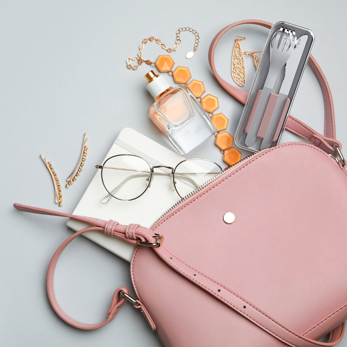 Estojo de talheres Monbento em aço inox, dentro de uma mala de senhora rosa, rodeado por acessórios femininos como óculos e perfume.