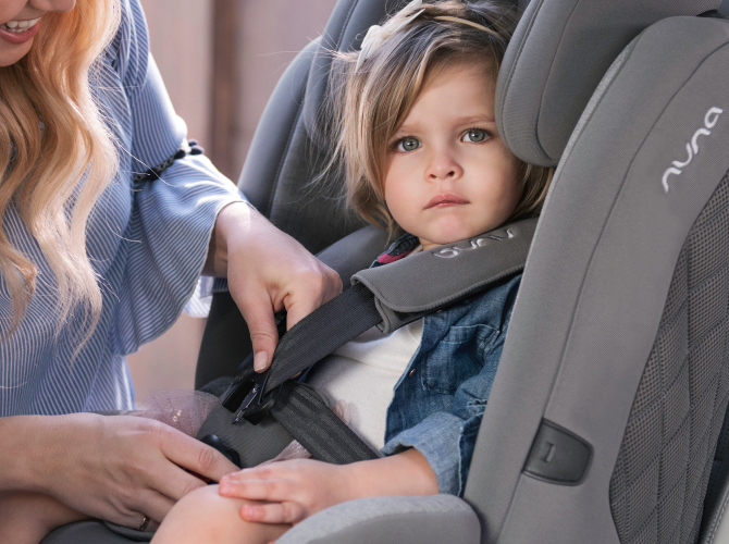 Criança sentada na cadeira auto Nuna Myti enquanto um adulto ajusta o arnês.