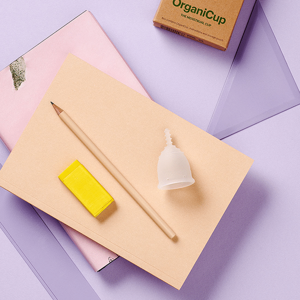 Copo menstrual OrganiCup Mini com material escolar sobre fundo lilás.