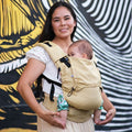 Mulher sorridente usando a Mochila Neko Switch com um bebé pequeno na frente, de pé em frente a um mural artístico.