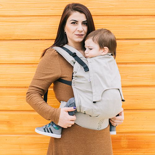 Mulher de cabelo castanho usando uma mochila porta-bebés Neko Ajustável Switch na cor Moss, carregando um bebé. A mochila é de cor cinza clara e ajusta-se ao corpo da criança.