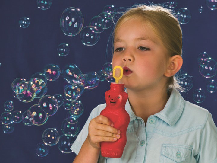 Uma menina soprando bolhas de sabão com o frasco em forma de urso vermelho da Pustefix. Bolhas grandes e brilhantes flutuam ao redor dela.