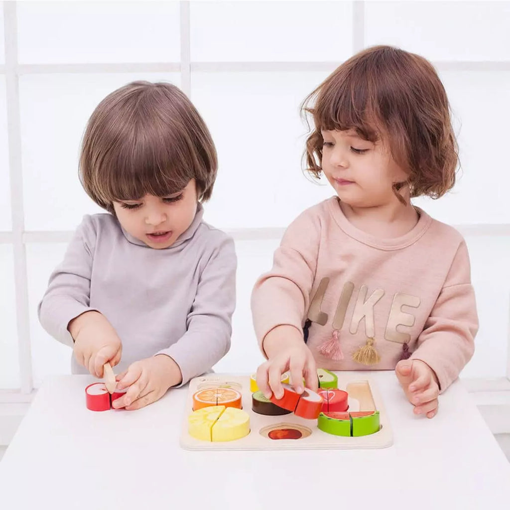 Duas crianças concentradas a brincar com o Classic World Puzzle de Cortar, uma a cortar uma maçã e a outra a encaixar peças na tábua.