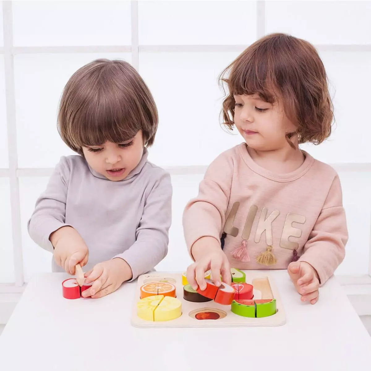 Duas crianças concentradas a brincar com o Classic World Puzzle de Cortar, uma a cortar uma maçã e a outra a encaixar peças na tábua.