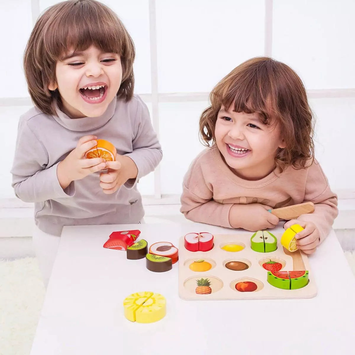 Duas crianças sorridentes brincam com o Classic World Puzzle de Cortar, com uma a segurar uma laranja e a outra a "cortar" uma pera na tábua de madeira.