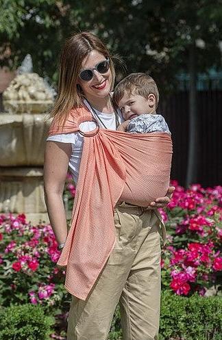 Mulher sorridente com óculos de sol, vestindo um sling de argolas Quokkababy Cool Air Pêssego e carregando um bebé adormecido, em fundo de jardim com flores coloridas.