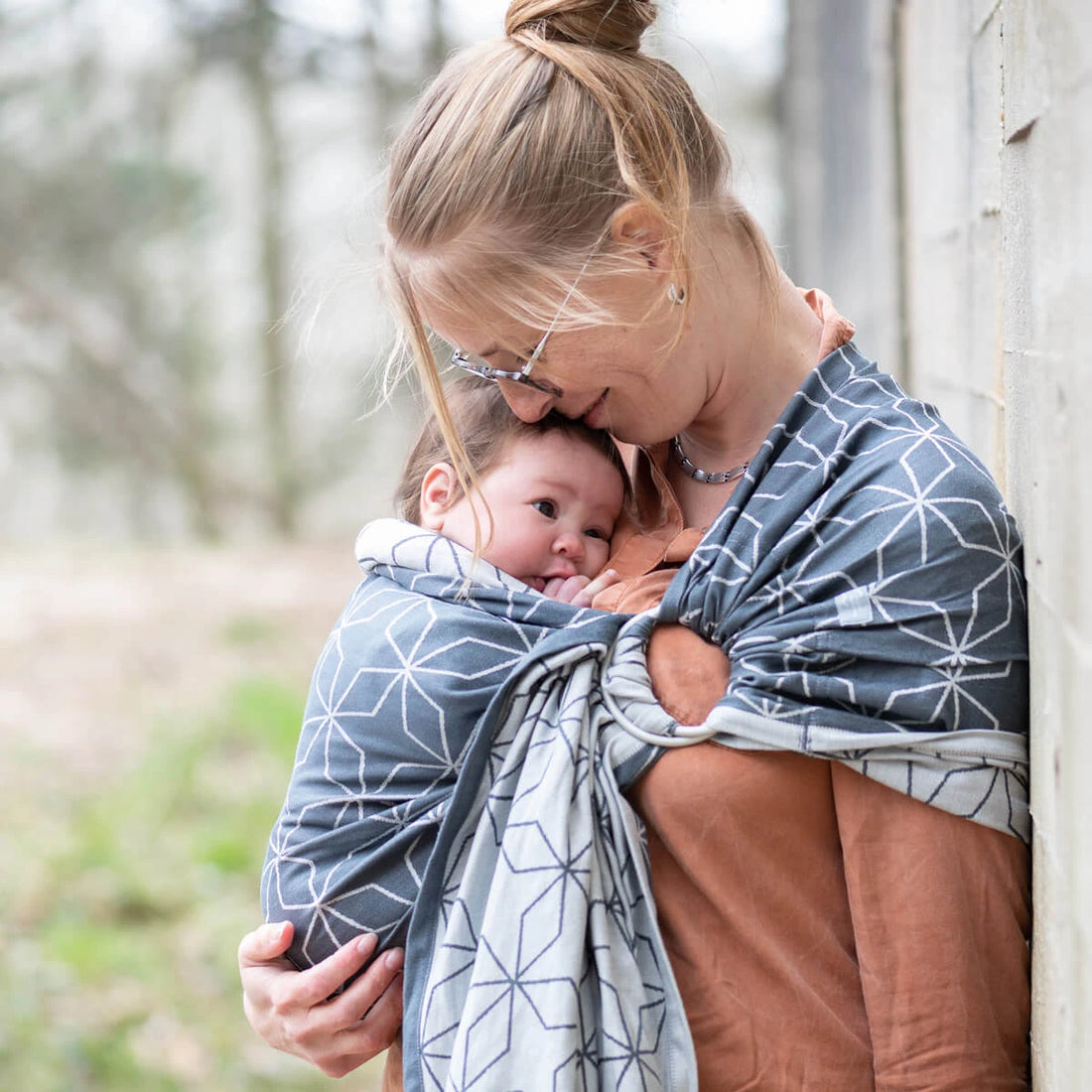 Uma mãe loira segura o seu bebé num sling de argolas Hoppediz Malmo Denim, sorrindo e abraçando-o carinhosamente. O sling é de tecido Jacquard azul e branco.