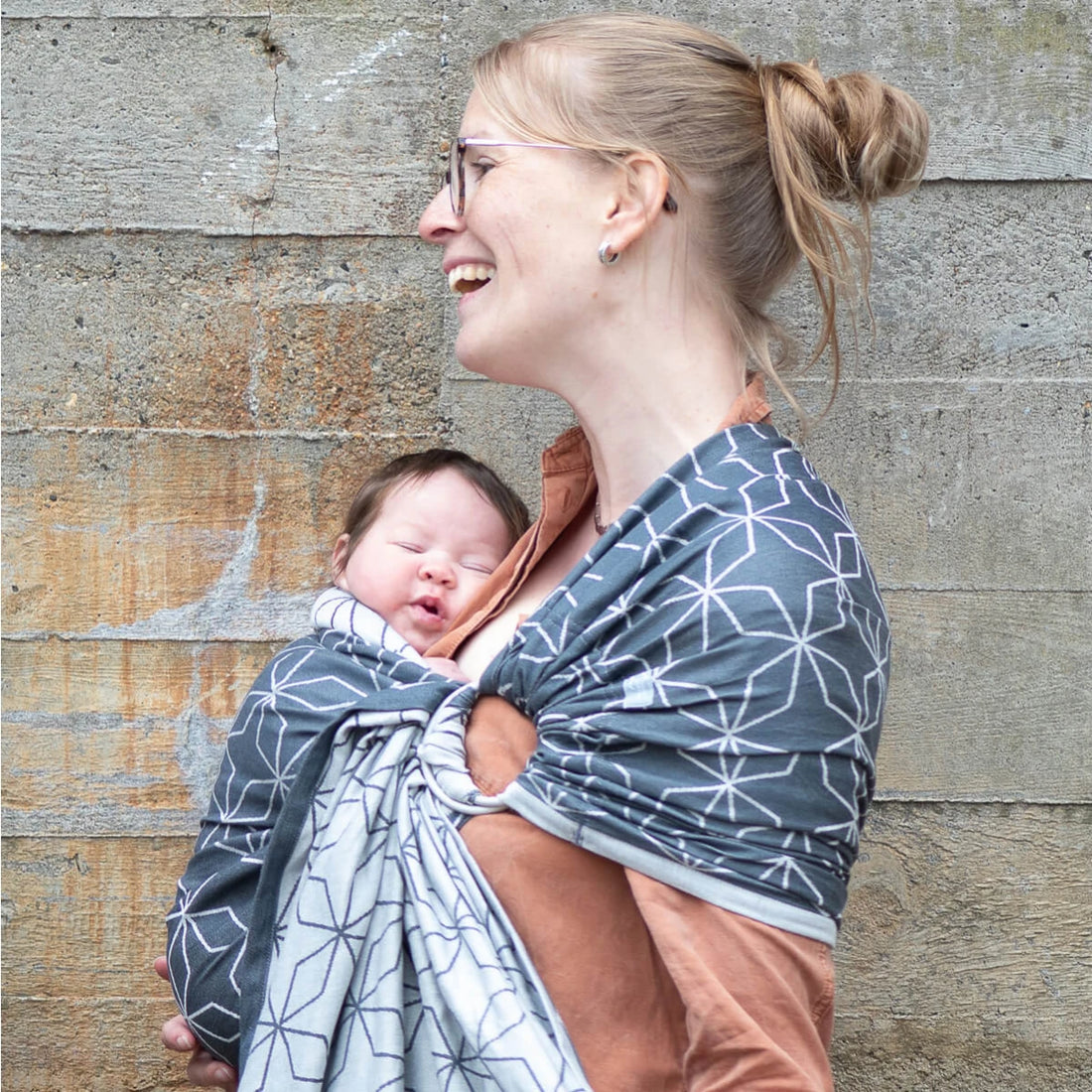 Uma mãe loira segura o seu bebé num sling de argolas Hoppediz Malmo Denim, sorrindo e abraçando-o carinhosamente. O sling é de tecido Jacquard azul e branco.