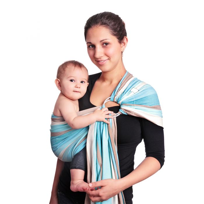 Uma mãe sorridente está a usar um sling de argolas Hoppediz Brest azul e bege, transportando um bebé pequeno no lado esquerdo do seu corpo. O sling está ajustado de forma a proporcionar suporte seguro e confortável ao bebé, mantendo-o numa posição ergonómica.