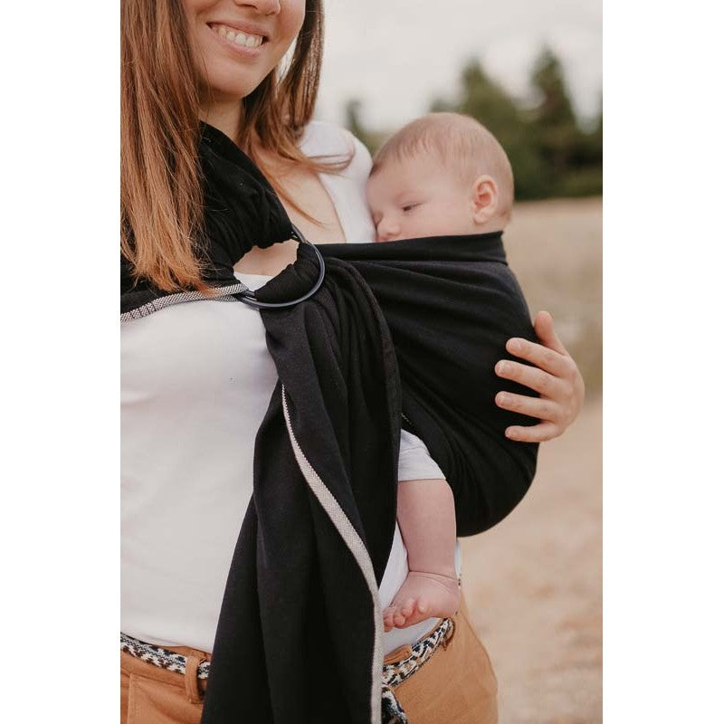 Mãe usando um sling de argolas preto Neobulle, segurando o seu bebé que está a dormir.