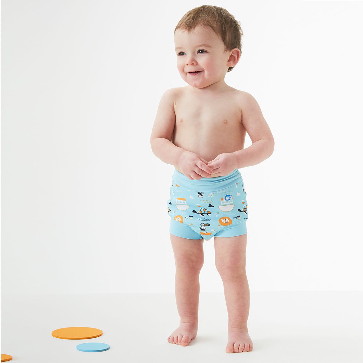 Bebé sorridente a usar a fralda de natação reutilizável Splash About Happy Nappy Noah's Ark, em azul claro com padrão.