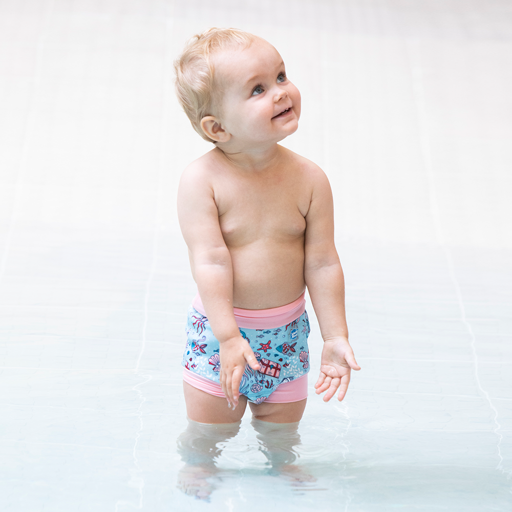 Bebé a usar fralda de natação Splash About Happy Nappy Hidden Treasure, em pé na água da piscina.
